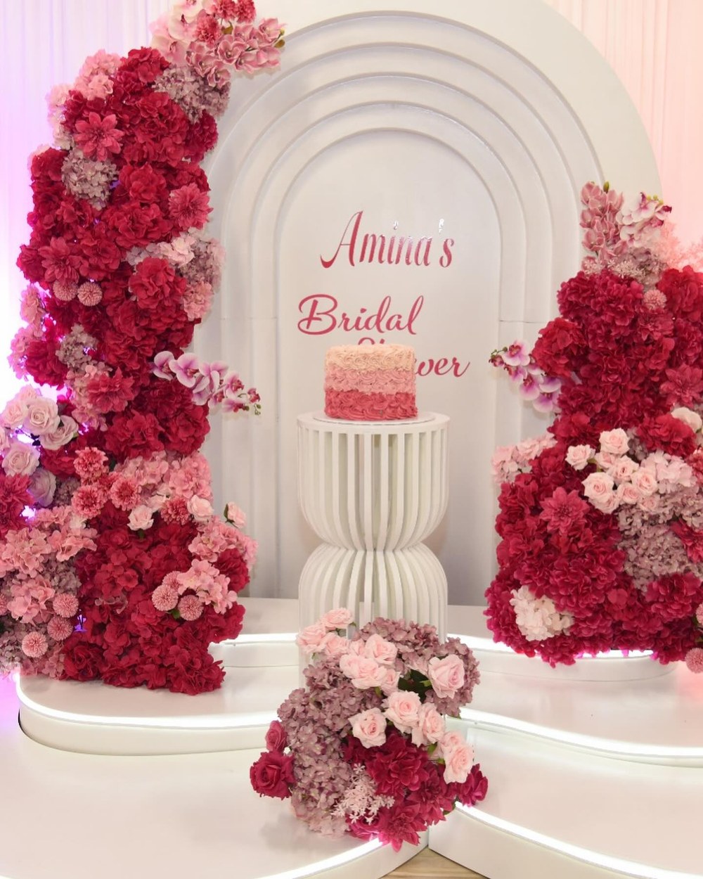 Bridal shower arch banked with deep pink and crimson roses around a cake plinth